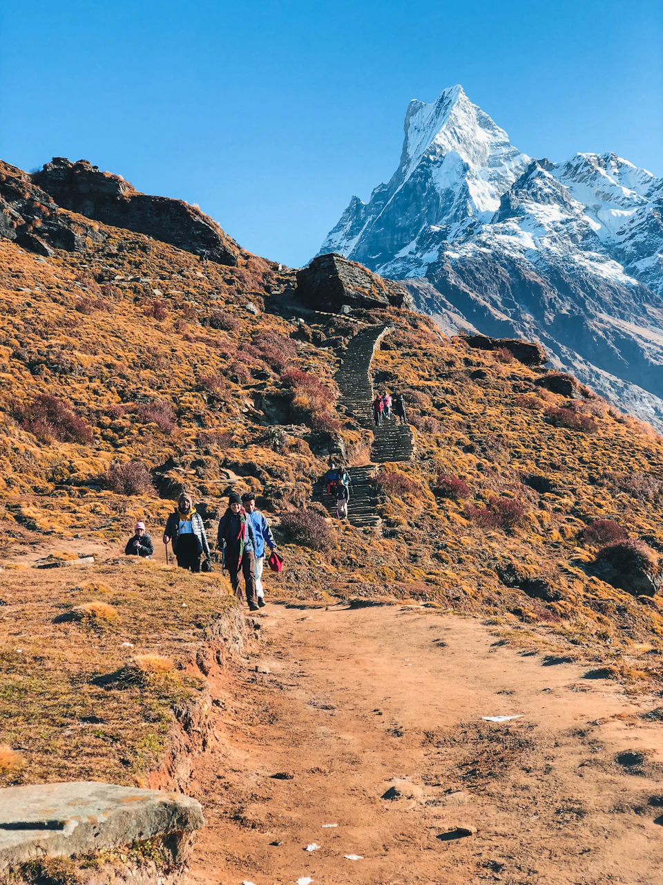 The image depicts a group of hikers ascending a mountain trail, surrounded by a rugged landscape with shrubs and rocks. The trail is marked by a set of stairs leading up to the peak, which is covered in snow. The sky above them is clear blue, suggesting a sunny day for their outdoor adventure. The hikers are seen walking along the trail, some carrying backpacks as they make their way towards the mountain's summit.