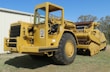A large, yellow Caterpillar construction vehicle with prominent wheels and heavy machinery components is positioned on grass beside a white metal building. The machine has visible levers and control mechanisms.