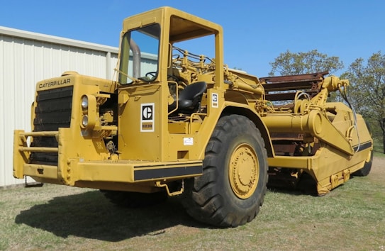 A large, yellow Caterpillar construction vehicle with prominent wheels and heavy machinery components is positioned on grass beside a white metal building. The machine has visible levers and control mechanisms.