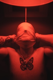 A person stands with their upper body exposed, their face obscured by a cloth pulled over their head. Tattoos are visible on the arms and chest, including a large butterfly design. The scene is bathed in a red light, creating an intense, dramatic atmosphere.