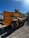 A large yellow JCB construction vehicle with a telescopic boom is parked outdoors on a gravel surface. It is surrounded by greenery and under a clear blue sky with sunlight casting shadows.