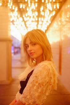 A person with blond hair stands in a softly lit hallway adorned with a ceiling of glowing overhead lights. The ambient light casts a warm, golden hue around her, creating a dreamy and ethereal atmosphere. She wears a lace outfit with delicate details.