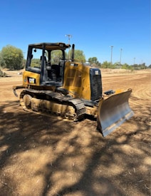 A yellow bulldozer is parked on a dirt surface under a clear blue sky. The machine has a Caterpillar logo and is equipped with tracks for movement and a large front blade for pushing material. In the background, there are sparse trees and some utility poles.