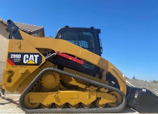 A yellow Caterpillar tracked loader with a robust design is parked outdoors, with a clear blue sky in the background. The machine features a label reading '299D XHP CAT' and is equipped with tracks for maneuverability on various terrains.