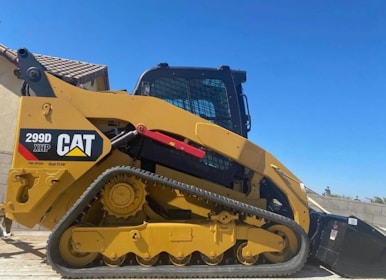 A yellow Caterpillar tracked loader with a robust design is parked outdoors, with a clear blue sky in the background. The machine features a label reading '299D XHP CAT' and is equipped with tracks for maneuverability on various terrains.