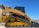 A yellow Caterpillar tracked loader with a robust design is parked outdoors, with a clear blue sky in the background. The machine features a label reading '299D XHP CAT' and is equipped with tracks for maneuverability on various terrains.