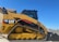 A yellow Caterpillar tracked loader with a robust design is parked outdoors, with a clear blue sky in the background. The machine features a label reading '299D XHP CAT' and is equipped with tracks for maneuverability on various terrains.