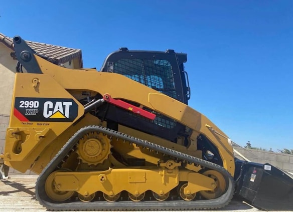 A yellow Caterpillar tracked loader with a robust design is parked outdoors, with a clear blue sky in the background. The machine features a label reading '299D XHP CAT' and is equipped with tracks for maneuverability on various terrains.