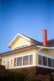 A yellow two-story house with white trim and a red brick chimney stands against a clear blue sky. The building features a gabled roof and a series of windows on the enclosed porch. It is elevated on a stone foundation, surrounded by green foliage at the base.