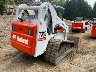 Multiple tracked skid steer loaders are parked on a dirt surface. The machinery, labeled with the brand name Bobcat and model T300, has a combination of white and bright red colors. The vehicles are surrounded by a natural setting, with trees in the background and construction materials nearby.