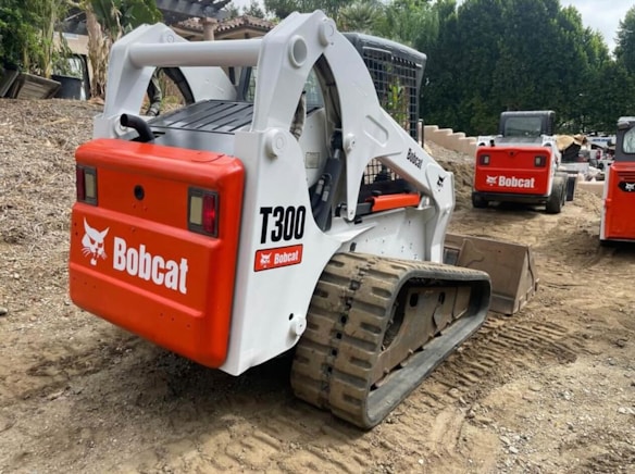 Multiple tracked skid steer loaders are parked on a dirt surface. The machinery, labeled with the brand name Bobcat and model T300, has a combination of white and bright red colors. The vehicles are surrounded by a natural setting, with trees in the background and construction materials nearby.