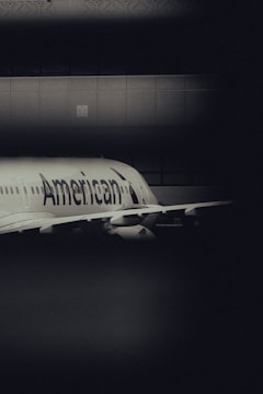 A dimly lit scene featuring a large aircraft with the name 'American' visible on the body. The plane is partially obscured by a dark foreground and set against a tiled wall with intricate patterns. The lighting creates a moody atmosphere with high contrast between light and shadow.