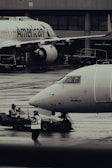 Two airplanes are stationed on a wet airport tarmac, with visible airline branding. Ground crew members in reflective vests are involved in operations, with one person directing and another operating equipment. The setting is industrial with concrete structures in the background.