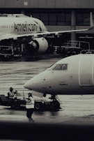 Two airplanes are stationed on a wet airport tarmac, with visible airline branding. Ground crew members in reflective vests are involved in operations, with one person directing and another operating equipment. The setting is industrial with concrete structures in the background.