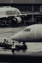 Two airplanes are stationed on a wet airport tarmac, with visible airline branding. Ground crew members in reflective vests are involved in operations, with one person directing and another operating equipment. The setting is industrial with concrete structures in the background.