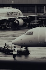Two airplanes are stationed on a wet airport tarmac, with visible airline branding. Ground crew members in reflective vests are involved in operations, with one person directing and another operating equipment. The setting is industrial with concrete structures in the background.