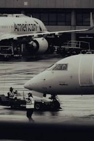 Two airplanes are stationed on a wet airport tarmac, with visible airline branding. Ground crew members in reflective vests are involved in operations, with one person directing and another operating equipment. The setting is industrial with concrete structures in the background.