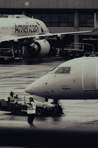 Two airplanes are stationed on a wet airport tarmac, with visible airline branding. Ground crew members in reflective vests are involved in operations, with one person directing and another operating equipment. The setting is industrial with concrete structures in the background.
