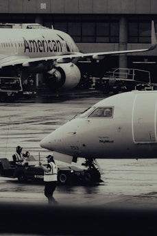 Two airplanes are stationed on a wet airport tarmac, with visible airline branding. Ground crew members in reflective vests are involved in operations, with one person directing and another operating equipment. The setting is industrial with concrete structures in the background.
