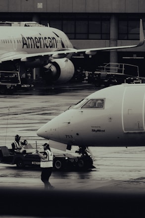 Two airplanes are stationed on a wet airport tarmac, with visible airline branding. Ground crew members in reflective vests are involved in operations, with one person directing and another operating equipment. The setting is industrial with concrete structures in the background.