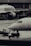 Two airplanes are stationed on a wet airport tarmac, with visible airline branding. Ground crew members in reflective vests are involved in operations, with one person directing and another operating equipment. The setting is industrial with concrete structures in the background.