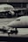 Two airplanes are stationed on a wet airport tarmac, with visible airline branding. Ground crew members in reflective vests are involved in operations, with one person directing and another operating equipment. The setting is industrial with concrete structures in the background.
