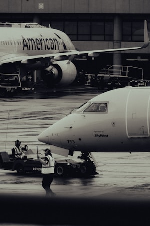 Two airplanes are stationed on a wet airport tarmac, with visible airline branding. Ground crew members in reflective vests are involved in operations, with one person directing and another operating equipment. The setting is industrial with concrete structures in the background.