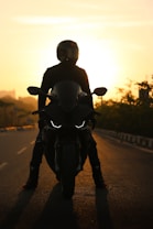 A silhouette of a person on a motorcycle is captured against a sunset backdrop. The sky is filled with warm hues of orange and yellow, casting a striking contrast against the dark figure at the center. The motorcycle is positioned on an empty road, and the rider is wearing a helmet, adding to the mysterious and adventurous aura.