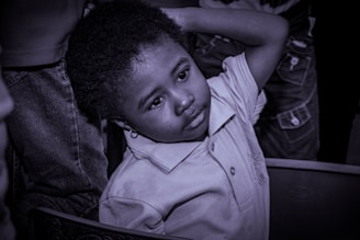 A young child with a thoughtful expression leans back slightly with one arm resting on their head. The child is wearing a collared shirt, and the scene is captured in a monochrome tone. Other people are partially visible in the background.