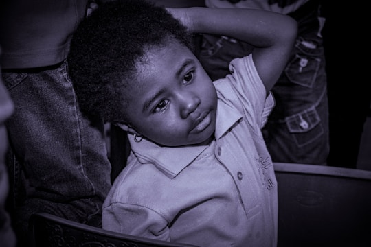 A young child with a thoughtful expression leans back slightly with one arm resting on their head. The child is wearing a collared shirt, and the scene is captured in a monochrome tone. Other people are partially visible in the background.