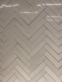 A herringbone pattern made of rectangular tiles with a glossy finish. The tiles are arranged in a diagonal zigzag layout, creating an intricate and visually appealing design. The surface reflects light, adding a sleek and modern feel.
