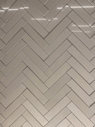 A herringbone pattern made of rectangular tiles with a glossy finish. The tiles are arranged in a diagonal zigzag layout, creating an intricate and visually appealing design. The surface reflects light, adding a sleek and modern feel.
