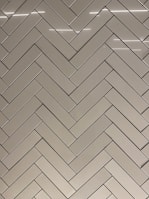 A herringbone pattern made of rectangular tiles with a glossy finish. The tiles are arranged in a diagonal zigzag layout, creating an intricate and visually appealing design. The surface reflects light, adding a sleek and modern feel.