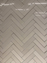 A herringbone pattern made of rectangular tiles with a glossy finish. The tiles are arranged in a diagonal zigzag layout, creating an intricate and visually appealing design. The surface reflects light, adding a sleek and modern feel.