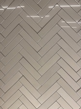 A herringbone pattern made of rectangular tiles with a glossy finish. The tiles are arranged in a diagonal zigzag layout, creating an intricate and visually appealing design. The surface reflects light, adding a sleek and modern feel.
