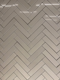 A herringbone pattern made of rectangular tiles with a glossy finish. The tiles are arranged in a diagonal zigzag layout, creating an intricate and visually appealing design. The surface reflects light, adding a sleek and modern feel.