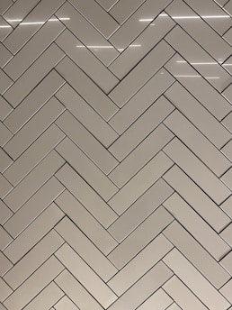 A herringbone pattern made of rectangular tiles with a glossy finish. The tiles are arranged in a diagonal zigzag layout, creating an intricate and visually appealing design. The surface reflects light, adding a sleek and modern feel.