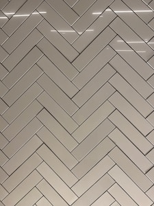 A herringbone pattern made of rectangular tiles with a glossy finish. The tiles are arranged in a diagonal zigzag layout, creating an intricate and visually appealing design. The surface reflects light, adding a sleek and modern feel.