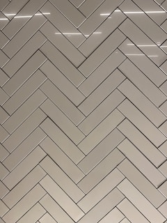 A herringbone pattern made of rectangular tiles with a glossy finish. The tiles are arranged in a diagonal zigzag layout, creating an intricate and visually appealing design. The surface reflects light, adding a sleek and modern feel.