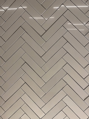 A herringbone pattern made of rectangular tiles with a glossy finish. The tiles are arranged in a diagonal zigzag layout, creating an intricate and visually appealing design. The surface reflects light, adding a sleek and modern feel.