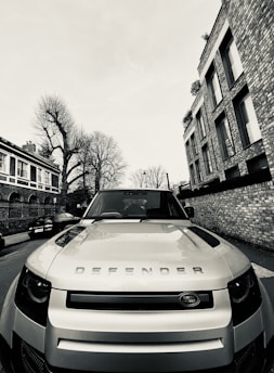 A Land Rover Defender is parked on the side of a street, positioned prominently in the foreground. Surrounding the vehicle are residential buildings with brick facades, and leafless trees lining the street. The image appears to have a monochromatic filter, enhancing the contrast between the car and the background.
