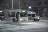 A trolleybus with a green and white color scheme is driving through a snowy urban street alongside a white van. The snow appears to be falling lightly, and the street is slightly covered. Buildings line the background, indicating a city setting, and the scene suggests nighttime with artificial lighting highlighting the vehicles.