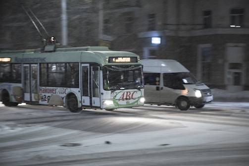 A trolleybus with a green and white color scheme is driving through a snowy urban street alongside a white van. The snow appears to be falling lightly, and the street is slightly covered. Buildings line the background, indicating a city setting, and the scene suggests nighttime with artificial lighting highlighting the vehicles.
