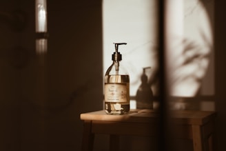 A glass bottle with a pump dispenser is placed on a wooden surface, illuminated by warm, natural light casting long shadows. The label on the bottle suggests it contains shea butter oil. The overall setting is minimalist, with a soft, earthy aesthetic.