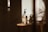A glass bottle with a pump dispenser is placed on a wooden surface, illuminated by warm, natural light casting long shadows. The label on the bottle suggests it contains shea butter oil. The overall setting is minimalist, with a soft, earthy aesthetic.
