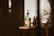 A glass bottle with a pump dispenser is placed on a wooden surface, illuminated by warm, natural light casting long shadows. The label on the bottle suggests it contains shea butter oil. The overall setting is minimalist, with a soft, earthy aesthetic.