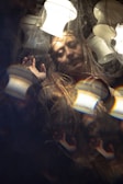 A person with a serene expression is framed within a surreal setting, surrounded by reflections and patterns. Soft lighting from multiple lamps casts gentle highlights across the scene, creating a dreamy and ethereal atmosphere. The person appears to be touching or pushing against a transparent barrier.