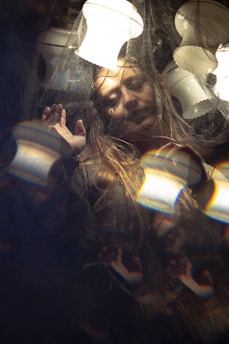 A person with a serene expression is framed within a surreal setting, surrounded by reflections and patterns. Soft lighting from multiple lamps casts gentle highlights across the scene, creating a dreamy and ethereal atmosphere. The person appears to be touching or pushing against a transparent barrier.
