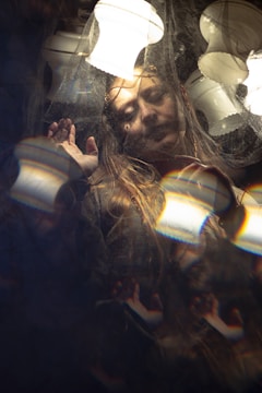 A person with a serene expression is framed within a surreal setting, surrounded by reflections and patterns. Soft lighting from multiple lamps casts gentle highlights across the scene, creating a dreamy and ethereal atmosphere. The person appears to be touching or pushing against a transparent barrier.