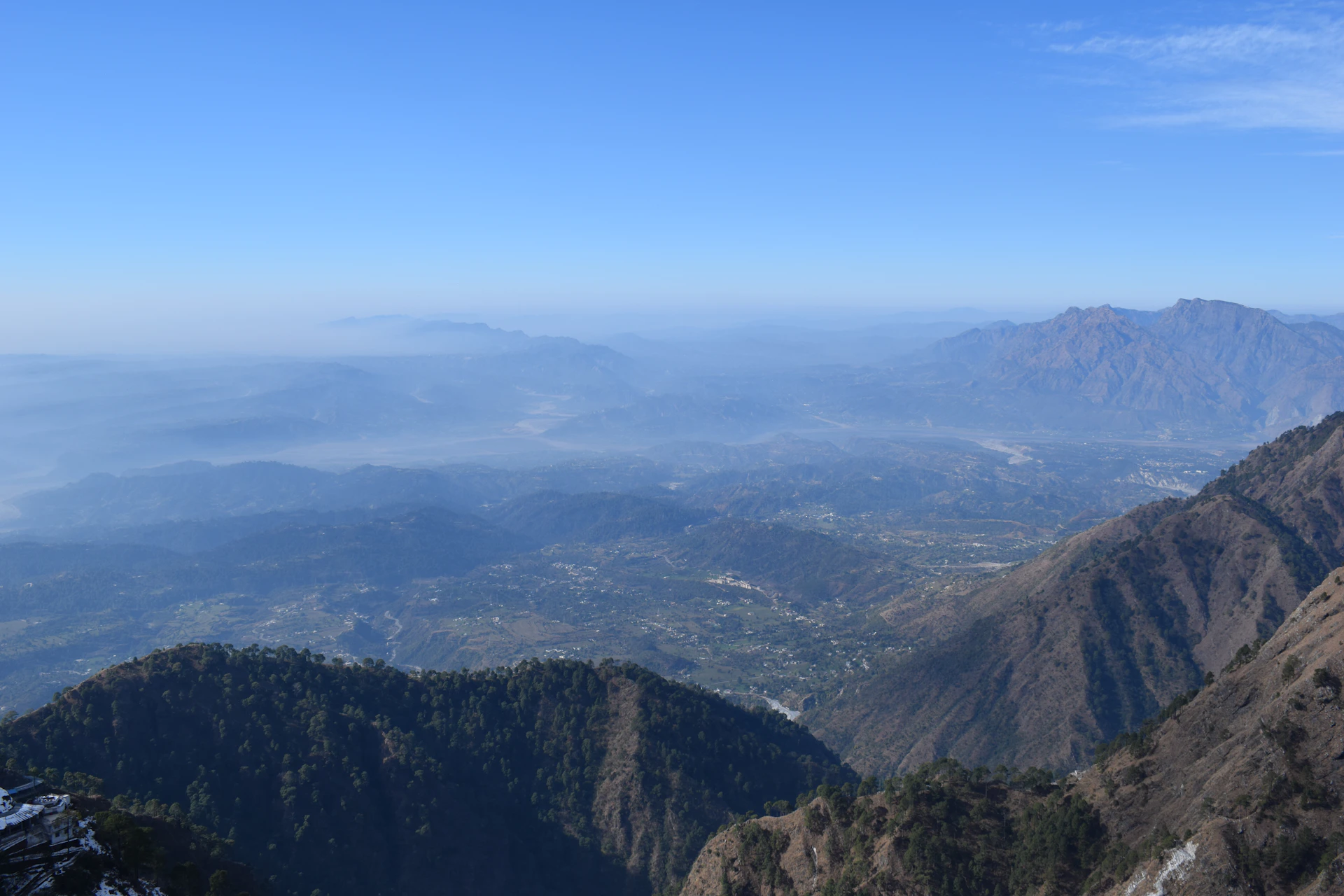 A panoramic view from the summit of Phulchoki, showing layers of forested hills stretching into the distance under a clear blue sky.